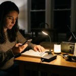 A woman uses a smartphone and reads a book by the light of an LED lamp and a power bank during a realistic power outage scenario, highlighting efficient emergency lighting and device charging.