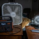 During a blackout, a large AC box fan struggles while plugged into a portable power station, indicating high drain, contrasted with a small, efficient 5V USB fan powered by a compact power bank, showing sustainable cooling.